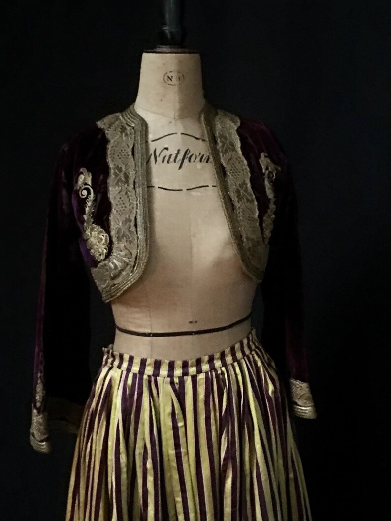 May include: A purple velvet jacket with gold embroidery and lace trim, paired with a yellow and purple striped skirt. The jacket has long sleeves and a cropped length. The skirt is full and has a high waist.