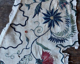 May include: Close-up of a section of embroidered fabric featuring floral and leaf designs. The embroidery uses shades of blue, green, and red on a cream-colored background. The fabric has a textured appearance with a frayed edge.