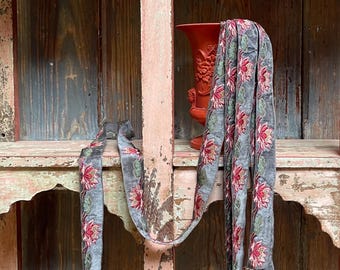 May include: A long, narrow grey fabric strip featuring a repeating embroidered pattern of pink lotus flowers with green leaves. The strip is draped over a tall, orange-red vase with a textured floral relief design on its exterior surface.