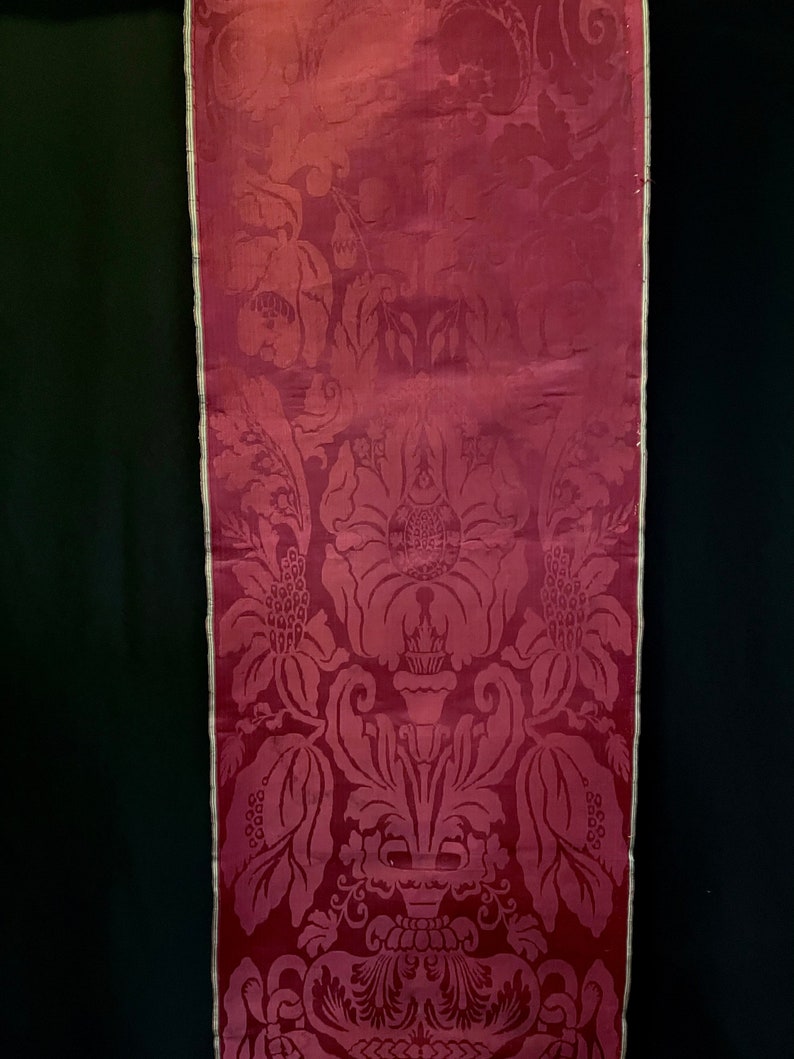 May include: A close-up of a deep red fabric with a damask pattern. The fabric has a subtle sheen and a delicate floral design.