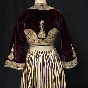 May include: A purple velvet jacket with gold embroidery and a matching striped skirt. The jacket has a high neckline and long sleeves with bell bottoms. The skirt is a full skirt with a high waist.