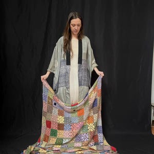 May include: A person wearing a gray and black patterned kimono holds a multi-colored crochet blanket. The blanket is made of squares in various colors, including red, green, blue, yellow, and pink.