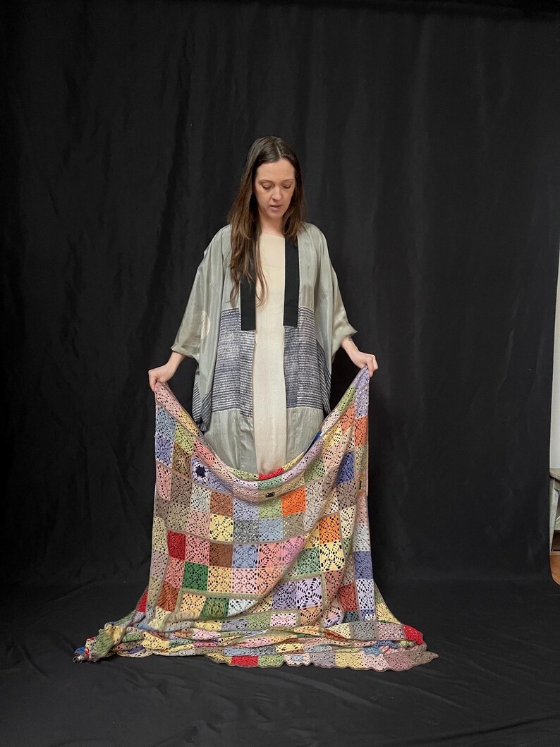 May include: A person wearing a gray and black patterned kimono holds a multi-colored crochet blanket. The blanket is made of squares in various colors, including red, green, blue, yellow, and pink.