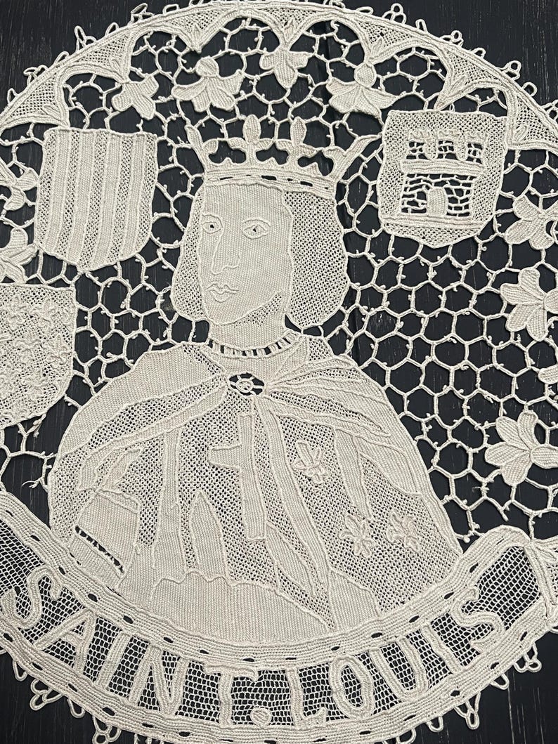 May include: Cream-colored lace doily featuring a portrait of a crowned figure, possibly a historical personage. The design includes heraldic shields, floral accents, and the text "SAINT LOUIS" in a banner. The intricate lace work is set against a dark background.