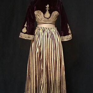 May include: A two-piece outfit featuring a burgundy velvet cropped jacket with gold embroidery and a yellow and burgundy striped skirt with a white lace trim.