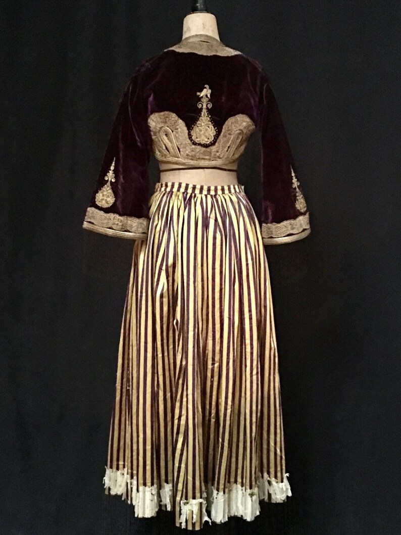 May include: A two-piece outfit featuring a burgundy velvet cropped jacket with gold embroidery and a yellow and burgundy striped skirt with a white lace trim.
