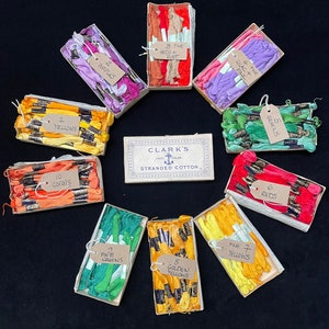 May include: Assortment of small boxes containing spools of embroidery floss in various colors, including yellow, purple, red, green, and coral. The boxes are labeled with the number of spools and the color name. The boxes are arranged in a circle around a larger box labeled "Clark's Fast Color Stranded Cotton."