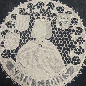 May include: Cream-colored lace doily featuring a portrait of Saint Louis, encircled by the text "SAINT LOUIS." The design includes a crowned figure, heraldic shields, and intricate floral and geometric patterns. The doily is round with a scalloped edge.