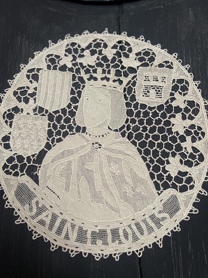 May include: Cream-colored lace doily featuring a portrait of Saint Louis, encircled by the text "SAINT LOUIS." The design includes a crowned figure, heraldic shields, and intricate floral and geometric patterns. The doily is round with a scalloped edge.