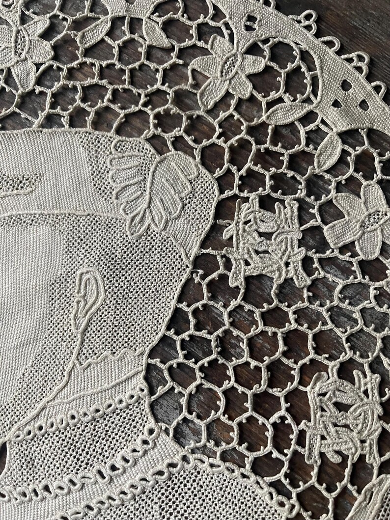 May include: Close-up of a beige lace doily with intricate floral and geometric patterns. The doily features a detailed design with flowers, leaves, and a delicate openwork mesh. The lace is finely crafted, showcasing the artistry of the textile.
