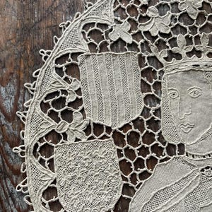 May include: Close-up of a beige, intricately crocheted doily. The design features a central portrait of a person wearing a crown, surrounded by floral and shield motifs. The doily's edge is scalloped, adding to its delicate appearance. The background is a dark, wooden surface.