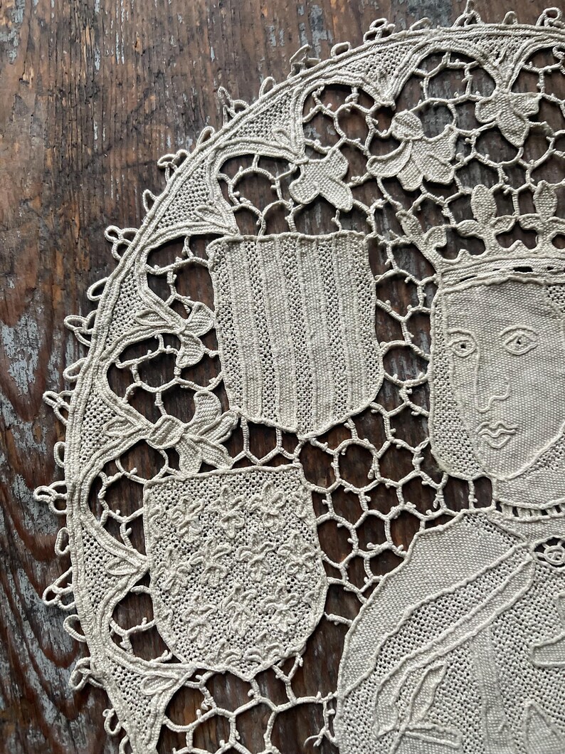 May include: Close-up of a beige, intricately crocheted doily. The design features a central portrait of a person wearing a crown, surrounded by floral and shield motifs. The doily's edge is scalloped, adding to its delicate appearance. The background is a dark, wooden surface.