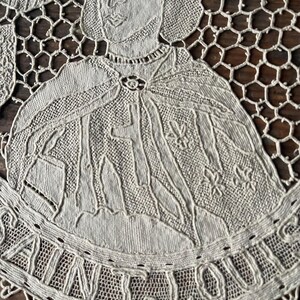 May include: Close-up of a beige lace doily featuring a detailed portrait of a historical figure, possibly a king or saint. The figure is wearing a cape and a crown. The word "SAINT LOUIS" is embroidered around the edge.