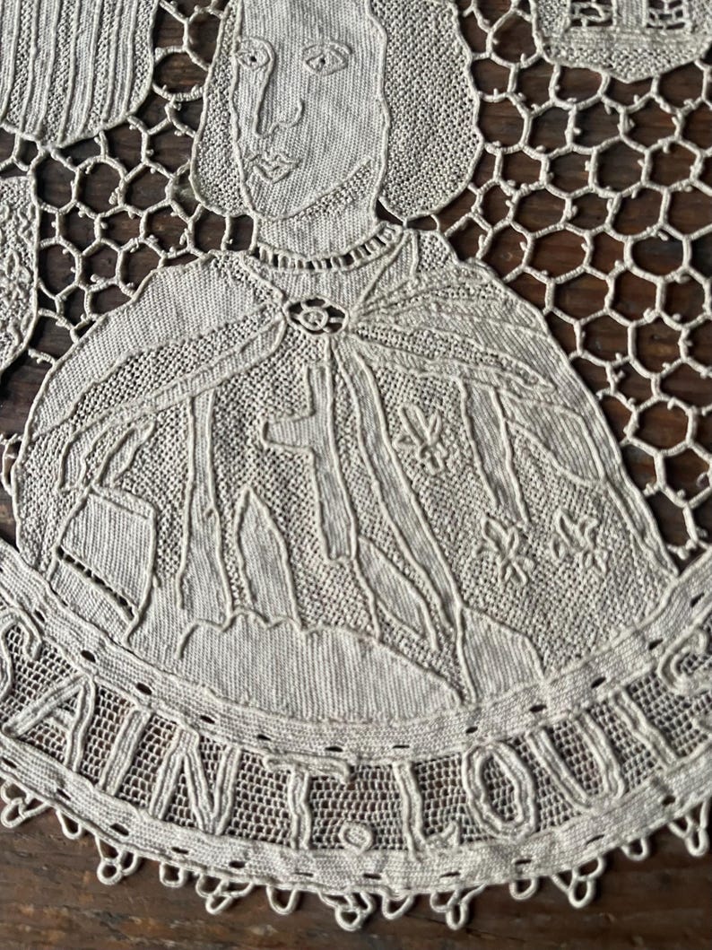 May include: Close-up of a beige lace doily featuring a detailed portrait of a historical figure, possibly a king or saint. The figure is wearing a cape and a crown. The word "SAINT LOUIS" is embroidered around the edge.