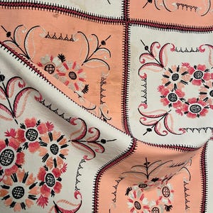 Puede incluir: Primer plano de una tela con un patrón floral repetido. El diseño presenta flores bordadas en tonos de rosa y blanco, sobre un fondo melocotón y blanco. Los bordes están delineados con un borde cosido de color rosa oscuro.