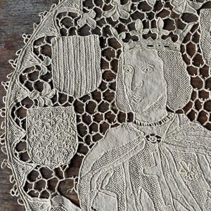 May include: Cream-colored, circular lace doily featuring a detailed portrait of a crowned figure, possibly royalty. The design includes two heraldic shields and intricate floral and geometric patterns. The doily is made of delicate, open-weave fabric.