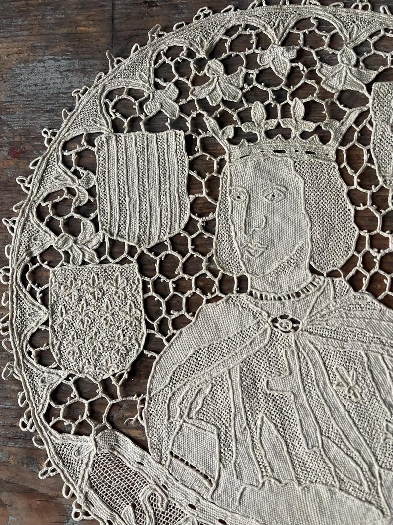 May include: Cream-colored, circular lace doily featuring a detailed portrait of a crowned figure, possibly royalty. The design includes two heraldic shields and intricate floral and geometric patterns. The doily is made of delicate, open-weave fabric.