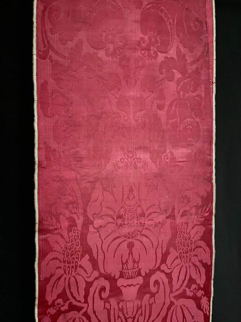 May include: A close-up of a red damask fabric with a floral pattern. The fabric has a white border and is woven with a raised design.