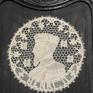 May include: Cream-colored, circular lace doily featuring a profile portrait of a man with a beard and the name "HENRI" in a banner. The lace has a floral border and a detailed openwork design.