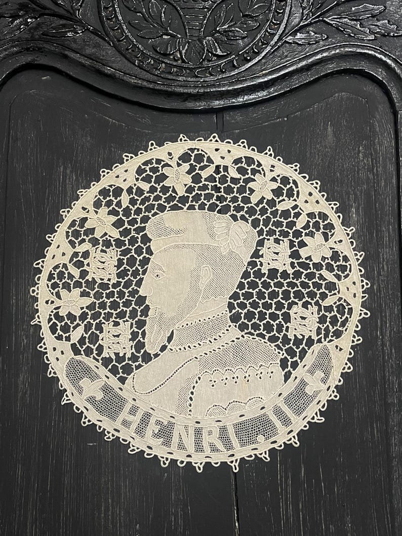 May include: Cream-colored, circular lace doily featuring a profile portrait of a man with a beard and the name "HENRI" in a banner. The lace has a floral border and a detailed openwork design.