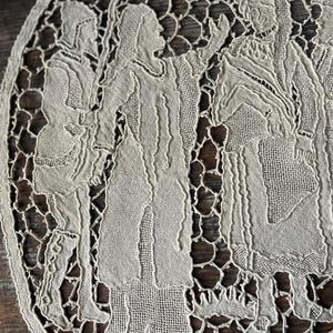 May include: An oval, off-white lace doily featuring a detailed depiction of figures in period clothing. The design includes multiple figures, with intricate cut-out patterns around the edges. The lace has a delicate, open weave.
