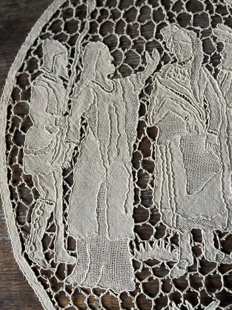 May include: An oval, off-white lace doily featuring a detailed depiction of figures in period clothing. The design includes multiple figures, with intricate cut-out patterns around the edges. The lace has a delicate, open weave.