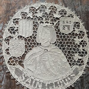 May include: A cream-colored, circular lace doily featuring a portrait of Saint Louis, with a crown and cape. The doily includes the text "SAINT LOUIS" and decorative shields. The lace has an open, intricate design.