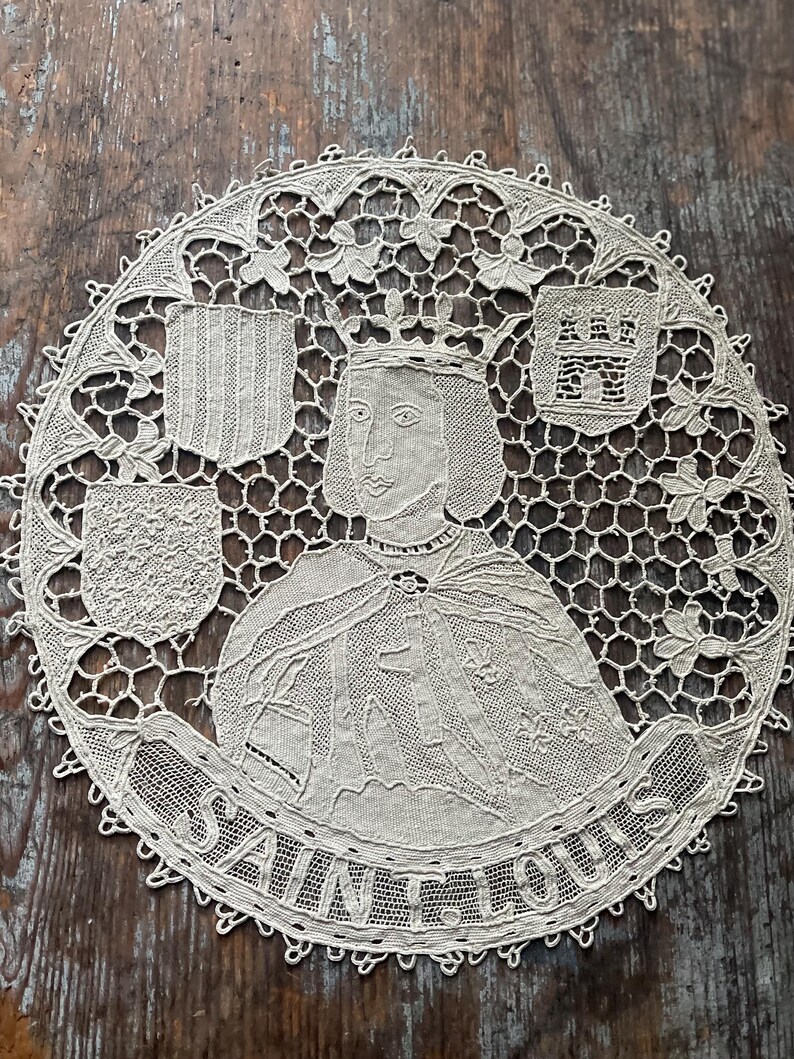 May include: A cream-colored, circular lace doily featuring a portrait of Saint Louis, with a crown and cape. The doily includes the text "SAINT LOUIS" and decorative shields. The lace has an open, intricate design.