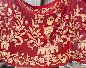 May include: A red textile with a repeating gold pattern featuring chalices, wheat stalks, and floral elements. The design includes a cross and the letters "2HL". The fabric is draped, showcasing the intricate details of the print.