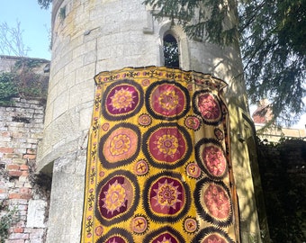 May include: A large, yellow, pink, and brown embroidered textile with a repeating pattern of circular medallions. The textile is hanging on a stone wall.