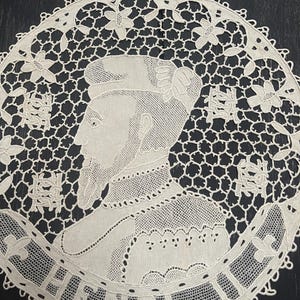 May include: A cream-colored, circular lace doily featuring a portrait of a man with the name "HENRI" in a banner. The doily has a detailed openwork design with floral and geometric patterns, and a scalloped edge.
