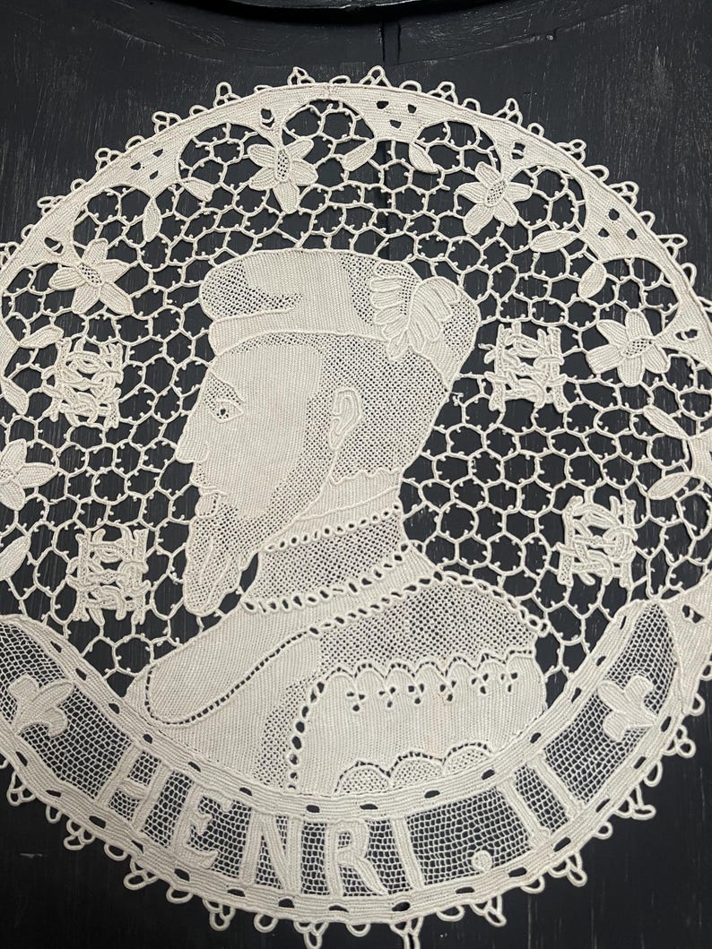 May include: A cream-colored, circular lace doily featuring a portrait of a man with the name "HENRI" in a banner. The doily has a detailed openwork design with floral and geometric patterns, and a scalloped edge.