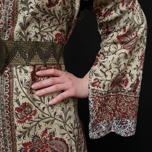 Pu&ograve; includere: Un abito a maniche lunghe color crema con stampa floreale rossa e nera. L'abito ha una cintura con perline e una vita drappeggiata.
