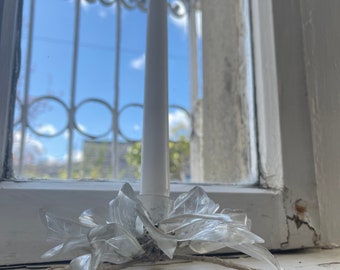 May include: A white taper candle in a clear, floral-shaped candle holder. The candle holder is made of clear plastic and sits on a windowsill. The background shows a blue sky and a decorative window.