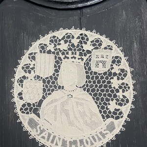 May include: A circular, cream-colored lace doily featuring a portrait of a crowned figure, possibly a king, with the text "SAINT LOUIS" around the image. The doily has a detailed, openwork design with floral and geometric patterns.