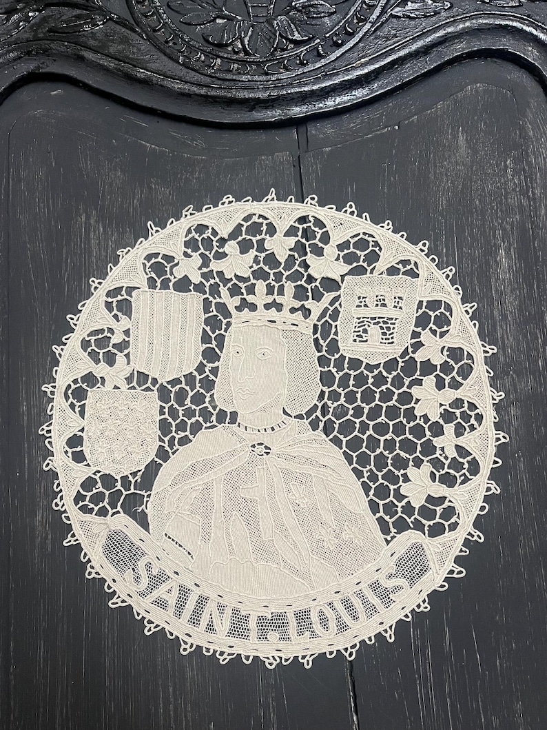 May include: A circular, cream-colored lace doily featuring a portrait of a crowned figure, possibly a king, with the text "SAINT LOUIS" around the image. The doily has a detailed, openwork design with floral and geometric patterns.
