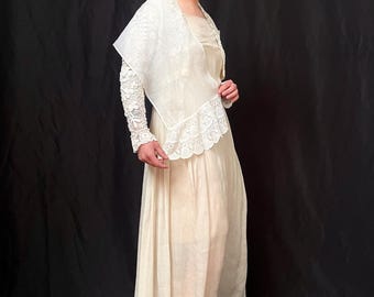 May include: A white maxi dress with a lace overlay and a white lace shawl. The dress has a scoop neckline and a flowing skirt. The model is wearing black wedge sandals.