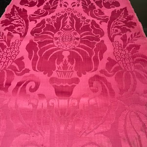 May include: A close-up of a pink fabric with a damask pattern. The pattern features a repeating design of stylized flowers and leaves.