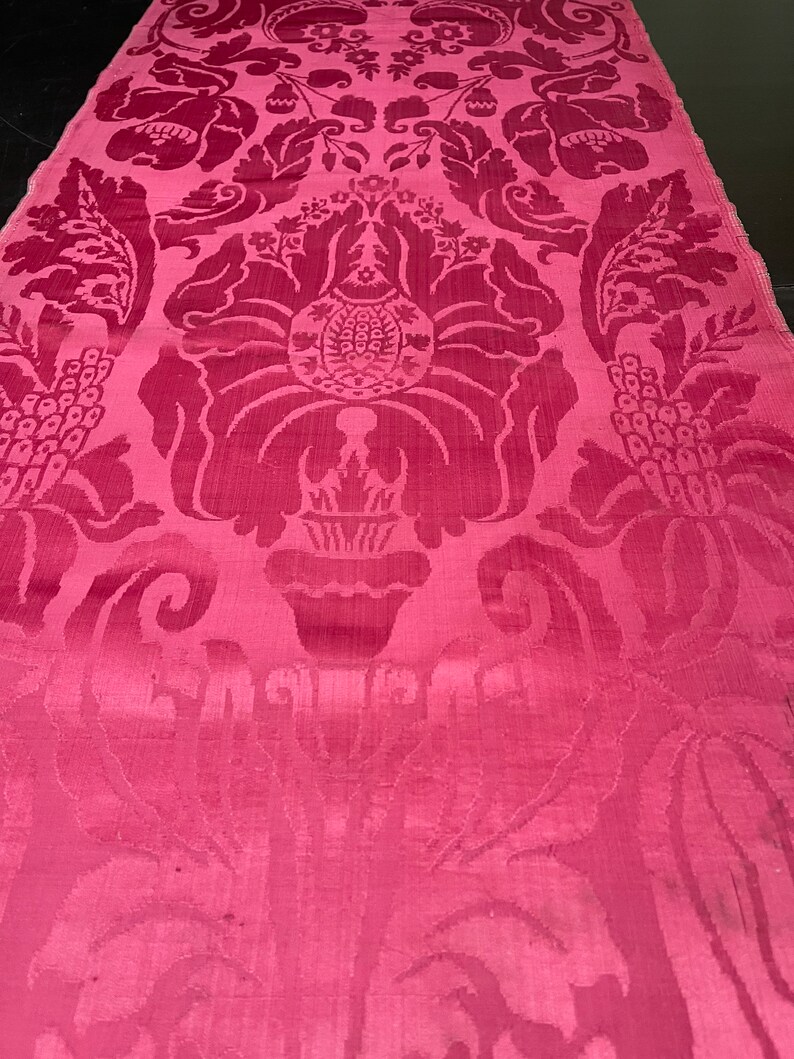 May include: A close-up of a pink fabric with a damask pattern. The pattern features a repeating design of stylized flowers and leaves.