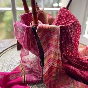 May include: A collection of pink and orange patterned fabric scarves with white and gold accents. The scarves are folded and arranged on a decorative metal tray.