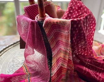 May include: A collection of pink and orange patterned fabric scarves with white and gold accents. The scarves are folded and arranged on a decorative metal tray.