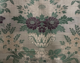 Può includere: Primo piano di un tessuto beige con un motivo ripetitivo di foglie verdi, fiori bianchi e bordeaux e un grappolo di uva bordeaux. Il tessuto ha un aspetto testurizzato e intrecciato.