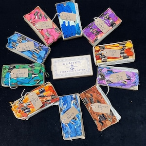 Könnte beinhalten: Eine Sammlung von 12 kleinen Schachteln mit Spulen von Stickgarn in verschiedenen Farben. Jede Schachtel ist mit einem Etikett versehen, das die Farbe und die Menge des Garns angibt. Die Schachteln sind in einem Kreis um eine größere Schachtel mit der Aufschrift "Clark's Fast Color Stranded Cotton" angeordnet.