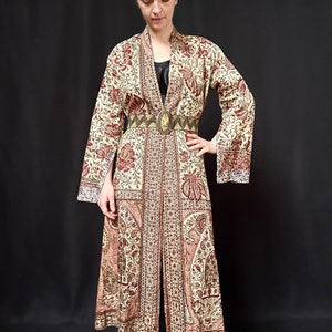 Pu&ograve; includere: Una lunga veste color crema con stampa floreale rossa e marrone. La veste ha una cintura con una grande fibbia decorata. La veste &egrave; realizzata in un tessuto morbido e fluido.