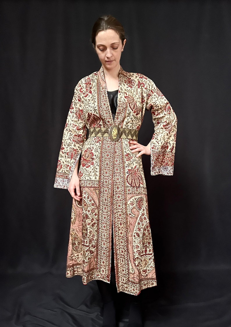Pu&ograve; includere: Una lunga veste color crema con stampa floreale rossa e marrone. La veste ha una cintura con una grande fibbia decorata. La veste &egrave; realizzata in un tessuto morbido e fluido.