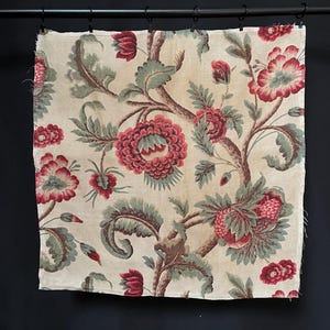 May include: A cream-colored fabric with a floral pattern. The flowers are red and green and have a vintage look. The fabric is hanging on a black rod.