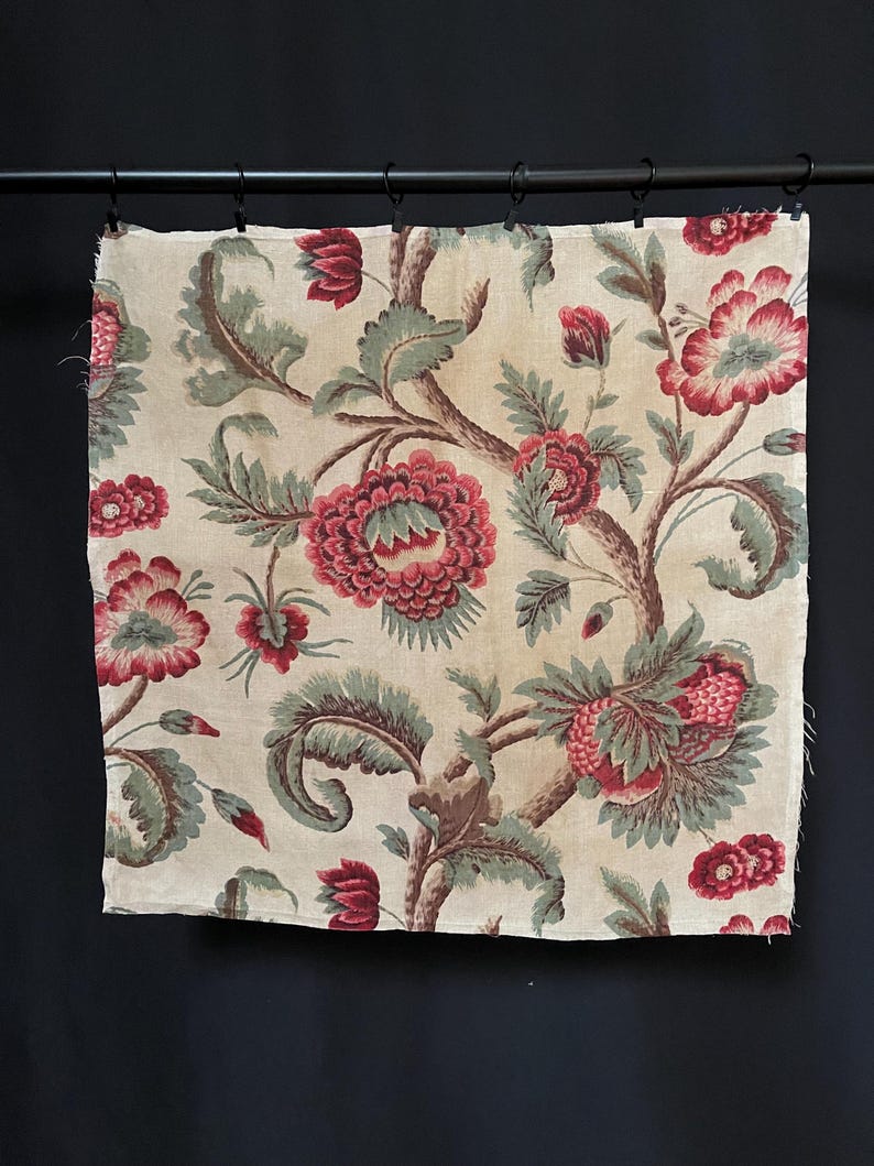 May include: A cream-colored fabric with a floral pattern. The flowers are red and green and have a vintage look. The fabric is hanging on a black rod.