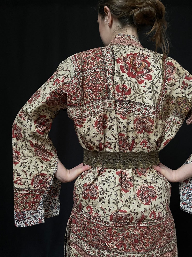 Pu&ograve; includere: Una lunga veste a maniche lunghe di colore crema con stampa floreale rossa e nera. La veste ha una silhouette ampia e fluida e una cintura con una fibbia dorata.