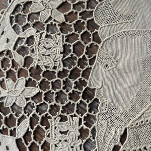 May include: Close-up of a beige lace doily with floral and geometric patterns. The doily features a detailed design with flowers, leaves, and a portrait of a woman. The lace is intricately woven, showcasing a delicate and vintage aesthetic.