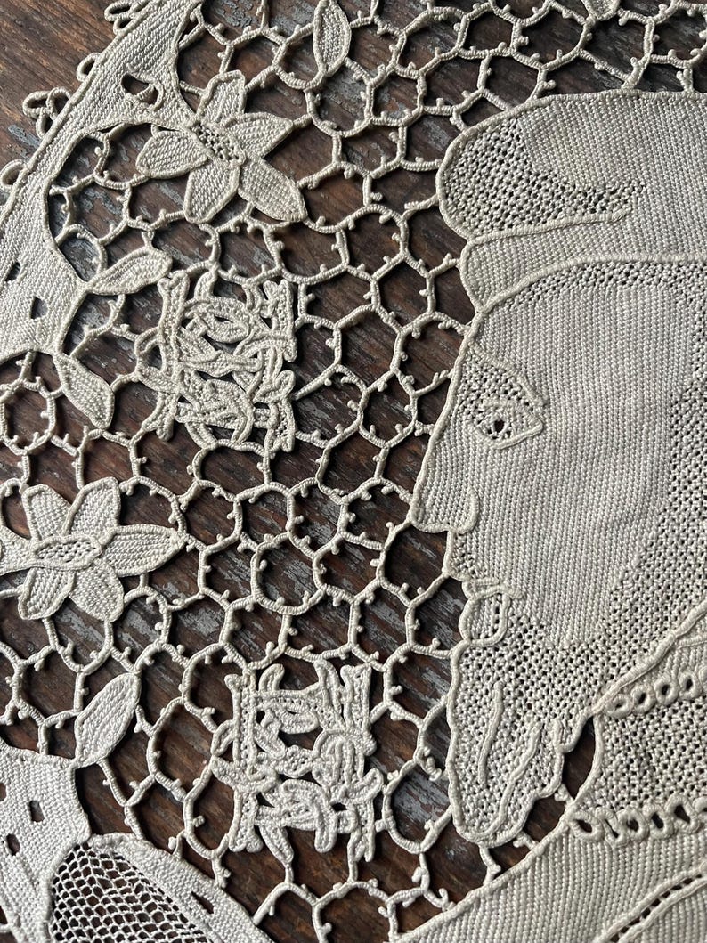 May include: Close-up of a beige lace doily with floral and geometric patterns. The doily features a detailed design with flowers, leaves, and a portrait of a woman. The lace is intricately woven, showcasing a delicate and vintage aesthetic.